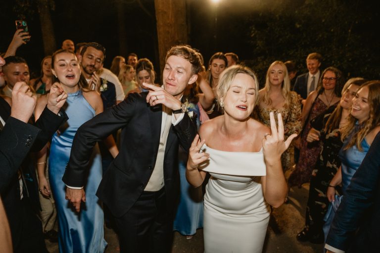 Magical evening moment as the bride and groom make their outdoor garden entrance at Tinakilly Country House in Ireland. With darkness settling in, additional lighting illuminates the couple while guests celebrate around them. This fine art wedding photograph captures romance, festive atmosphere, and the dramatic beauty of an evening celebration. Photographed by WeddingWithMe, it showcases professional wedding photography and videography, ideal for Irish wedding galleries, social media, and SEO-optimized luxury wedding website content.